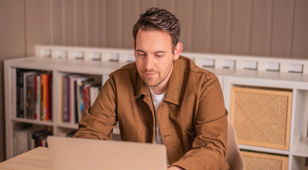 Bence Székely, en av gründerne av Frem Digital, sitter ved et skrivebord og ser på en laptop. I bakgrunnen står hyller med bøker og esker.