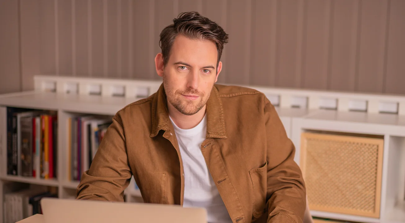 Bence Székely, Strategy Lead i Frem Digital, sitter ved et skrivebord med en laptop og ser inn i kameraet. I bakgrunnen står hyller med bøker og dekor.